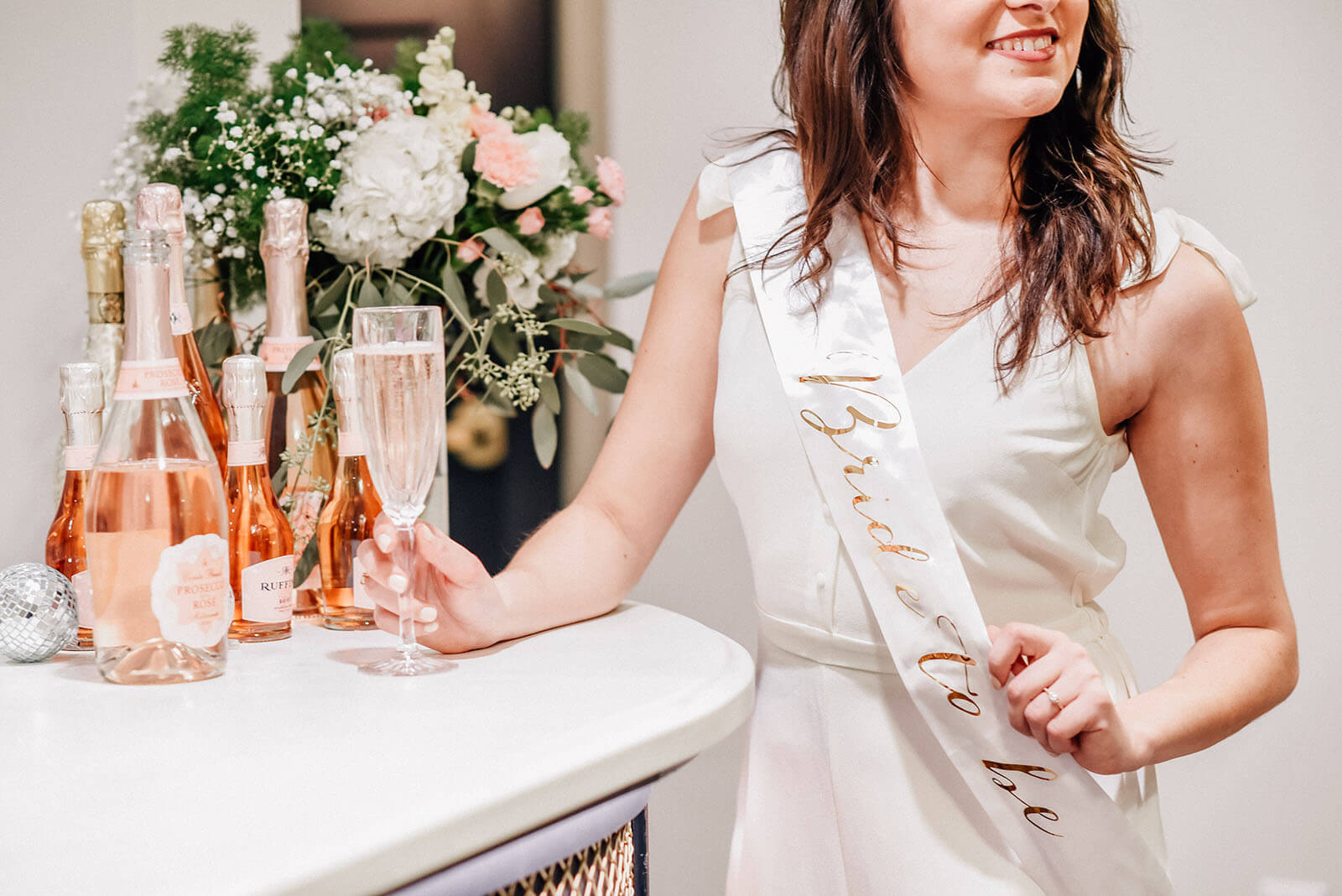 Woman wearing a 'bride to be' sash holding a glass of champagne leaning on the concession stand bar